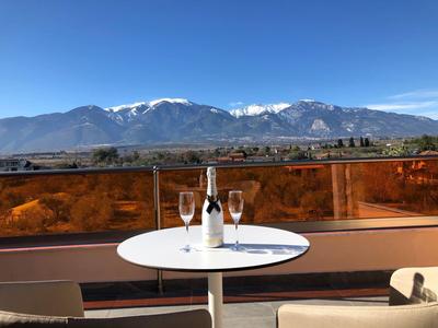 Runder weißer Tisch mit Weinflasche, Gläsern auf Balkon, dahinter Berge mit Schneespitzen, blauer Himmel.