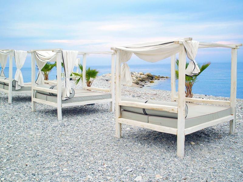 Weiße Strandbetten mit Vorhängen stehen auf Kieselstrand vor blauem Meer und Wolkenhimmel.