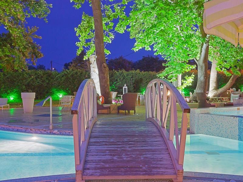 Holzbrücke über beleuchtetes Poolbecken, umgeben von grünen Bäumen und lauschiger Abendstimmung.