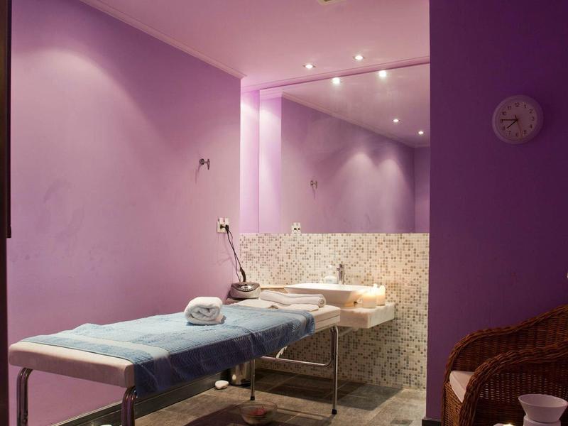 Ein lila Behandlungsraum mit Liege, großem Spiegel und Korbstuhl. Boden aus grauen Fliesen.