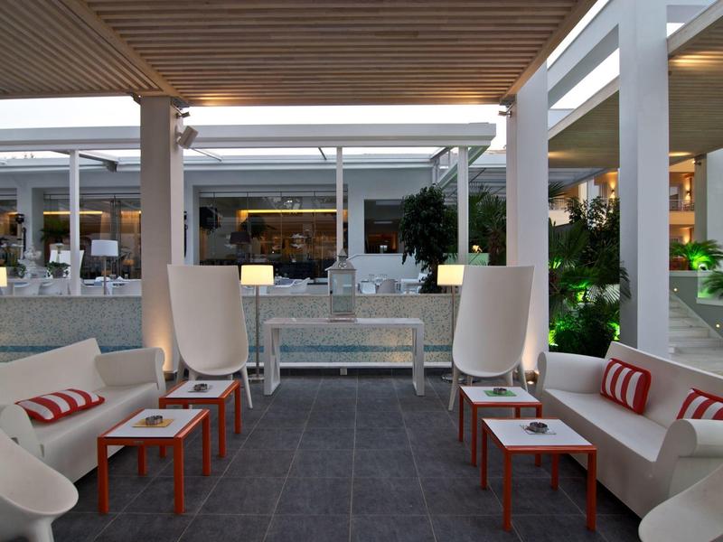 Moderne Lounge mit weißen Sesseln, roten Tischen und grauem Boden unter Holzdecke.