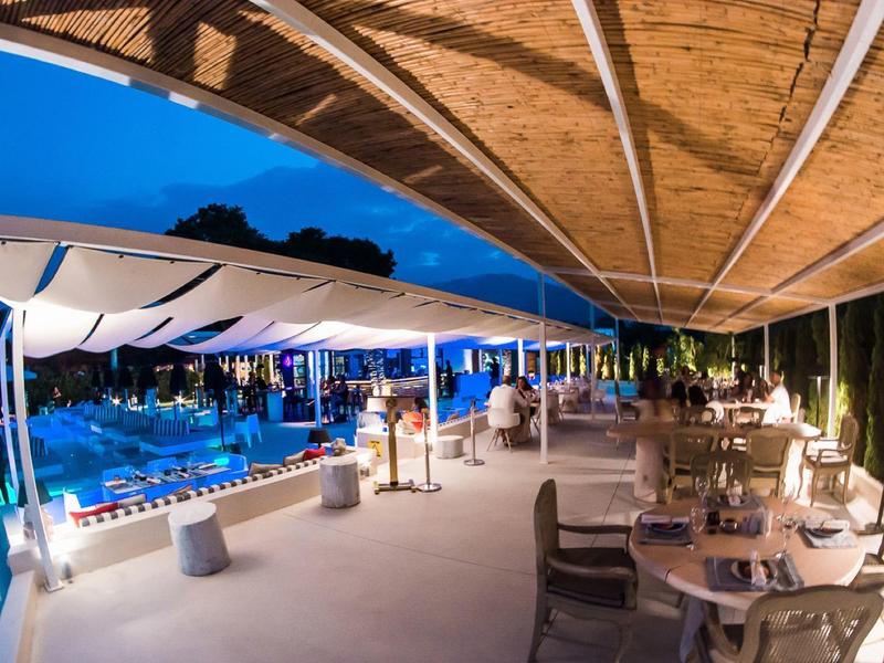 Eine beleuchtete Terrasse mit Tischen und Stühlen neben einem Pool unter einem Holz- und Stoffdach nachts.