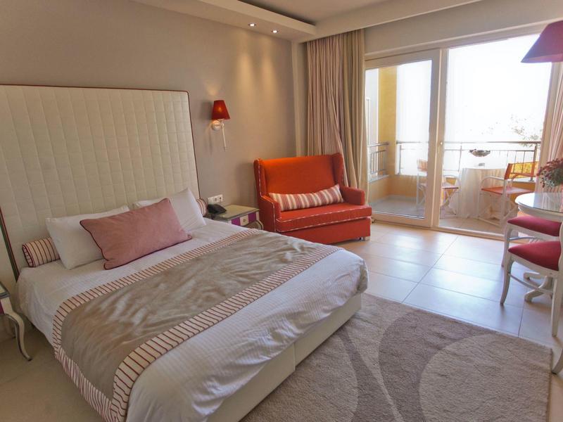 Hell eingerichtetes Schlafzimmer mit großem Bett, orangem Sofa und Balkon mit Glas-Schiebetür.