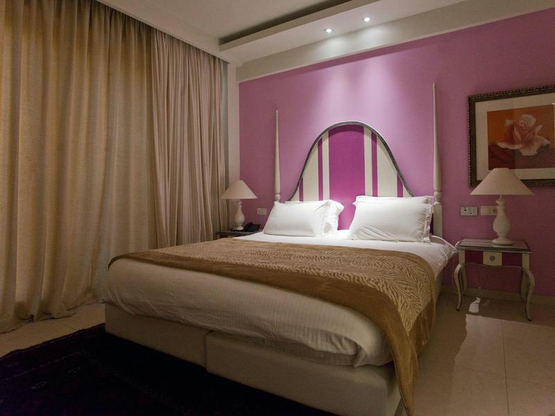 Schlafzimmer mit lila Wand, großem Bett, beigen Vorhängen und Nachttischen mit Lampen.