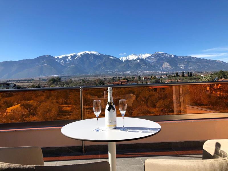 Runder weißer Tisch mit Weinflasche, Gläsern auf Balkon, dahinter Berge mit Schneespitzen, blauer Himmel.
