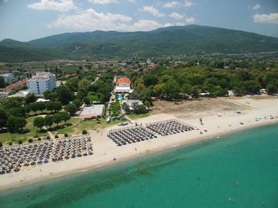 Luftaufnahme eines Strandes mit Sonnenschirmen, angrenzendem Hotel und grünen Hügeln im Hintergrund.