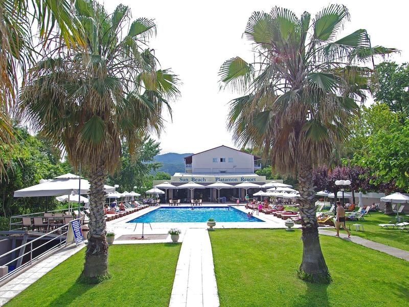 Blick auf einen Hotelpool mit Sonnenschirmen, Palmen und einem Gebäude im Hintergrund.