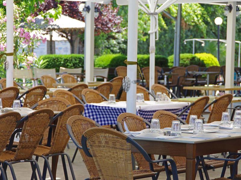 Freiluftrestaurant mit geflochtenen Stühlen und blau-weiß karierten Tischdecken unter Sonnenschirmen.