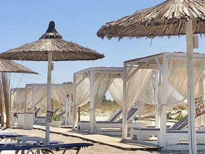 Strand mit Strohdach-Sonnenschirmen und weißen, halbtransparenten Vorhängen an Liegen.