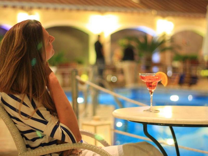 Frau sitzt entspannt am Pool mit Cocktail auf rundem Tisch, warme Beleuchtung, Abendstimmung.