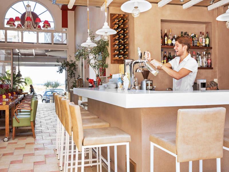 Helle Bar mit Holzstühlen, stehendem Barkeeper und großen Fenstern, durch die Tageslicht strömt.