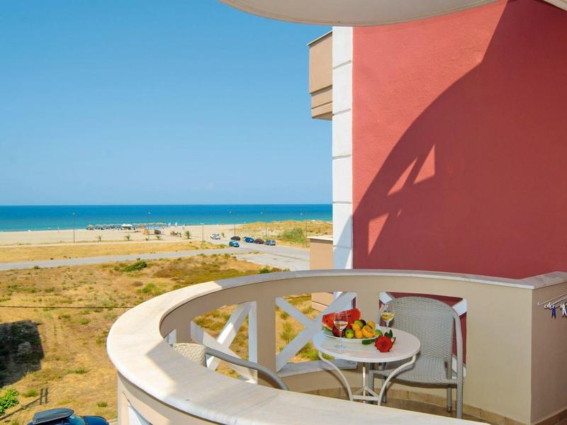 Balkon mit weißem Geländer, Tisch mit Wasser und Früchten, Sandstrand und blaues Meer im Hintergrund.