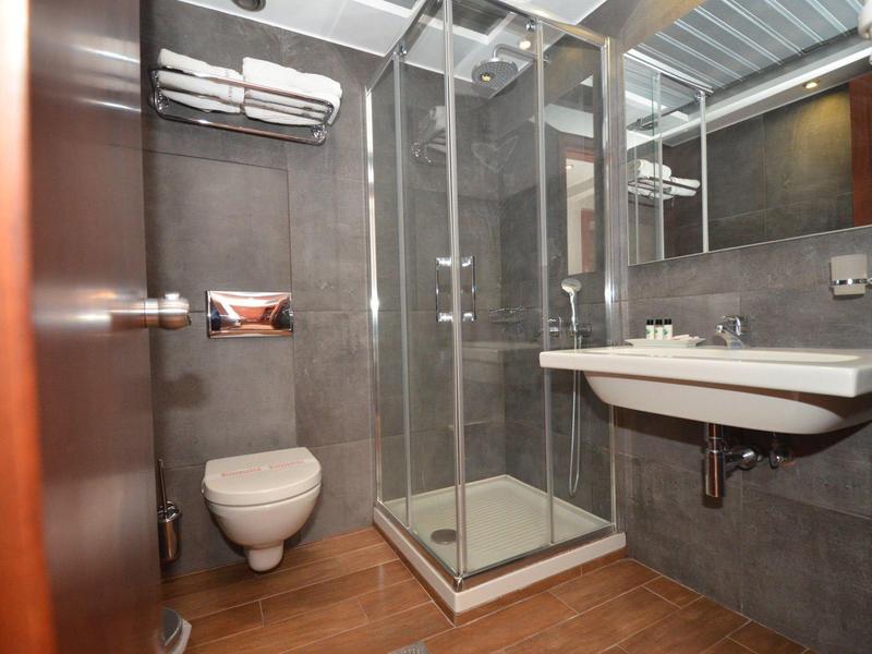 Kleines Badezimmer mit Glasdusche, Wand-WC, weißem Waschbecken und grauen Wandfliesen.