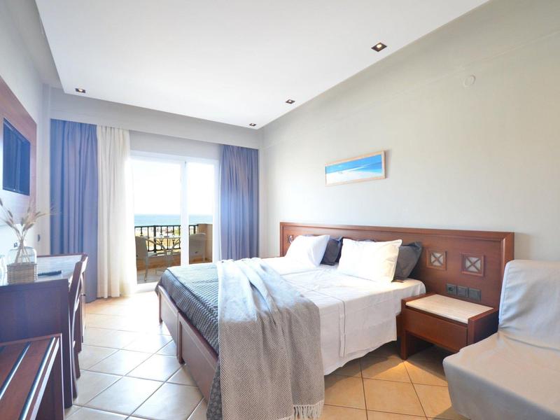 Helles Hotelzimmer mit Doppelbett, Holzmöbeln, hellen Fliesen und Balkon im Hintergrund.