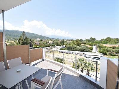 Balkon mit Tisch und Stühlen, blickt auf grüne Landschaft und Straßen unter blauem Himmel.