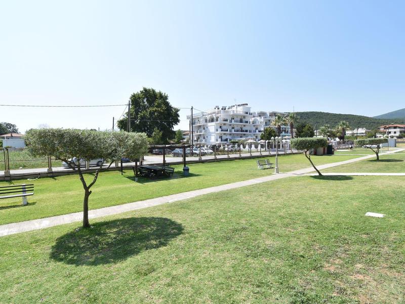 Grüner Park mit gepflegtem Rasen, einzelnen Bäumen, Weg und Gebäuden im Hintergrund unter blauem Himmel.