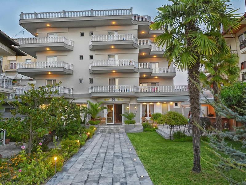 Mehrstöckiges Gebäude mit Balkonen, heller Fassade und Kiesweg, umgeben von grünen Pflanzen und Palmen.