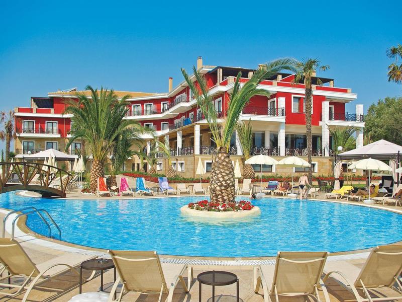 Großes, rundes Schwimmbecken vor mehrstöckigem Hotel mit roten Wänden und Palmen bei blauem Himmel.