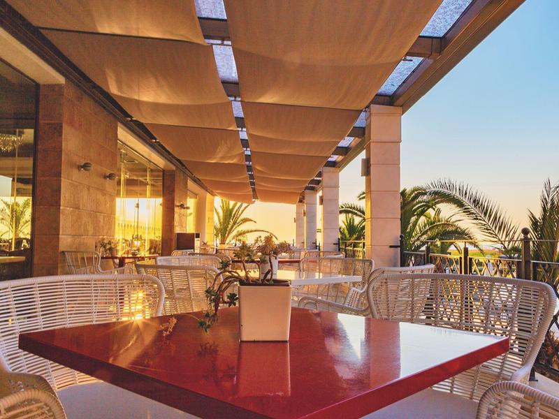 Terrasse mit rötlichem Tisch, weißen Stühlen, Holzdach und Blick auf Palmen bei Sonnenuntergang.