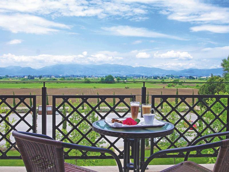 Balkon mit Stühlen, Tisch, Frühstück, Blick auf grüne Felder und Berge unter blauem Himmel.