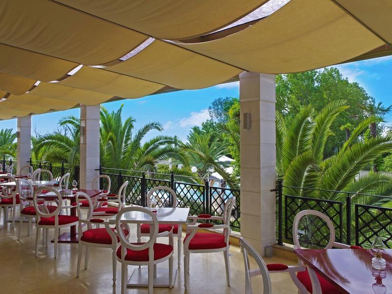 Überdachte Terrasse mit weißen Stühlen, roten Sitzkissen und Blick auf Palmen und blauen Himmel.