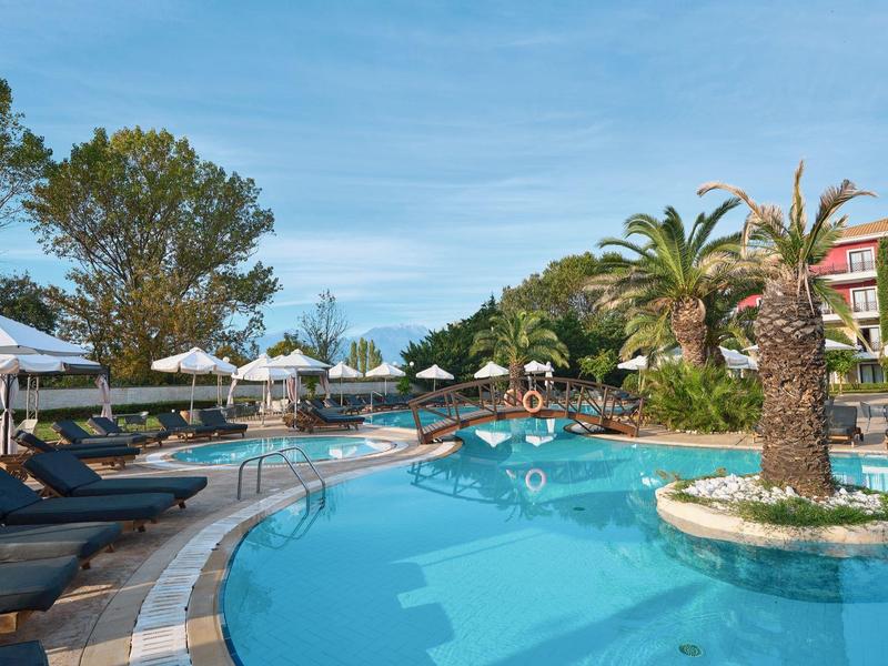 Freizeitbad mit klarem blauen Pool, Palmen, Sonnenliegen und hellem Sonnenschirm unter blauem Himmel.