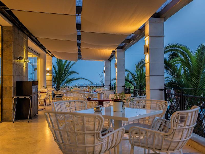 Offene Terrasse mit Glasgeländer, Rattanmöbeln und Palmen im Dämmerlicht.