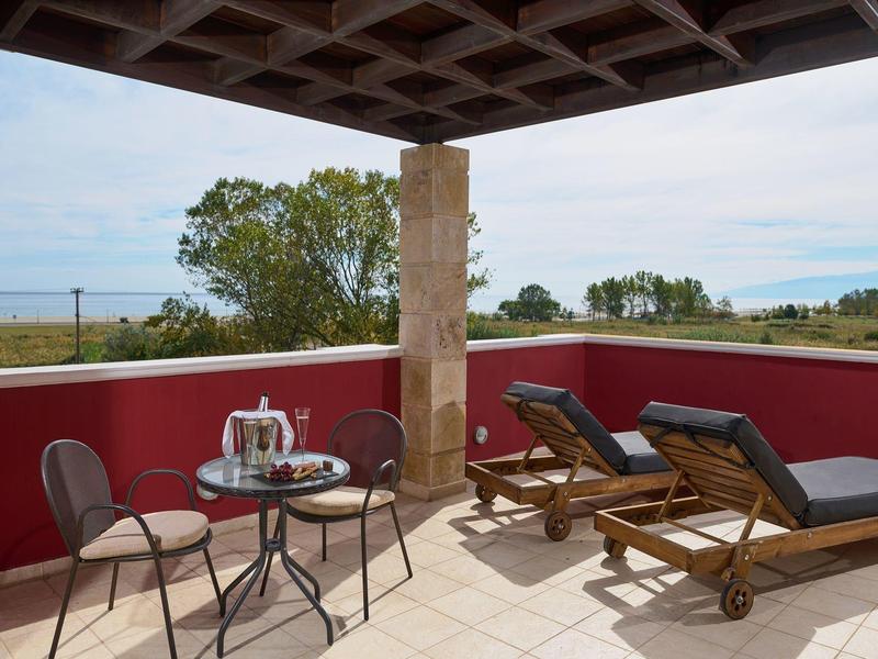 Terrasse mit roten Wänden, zwei Liegestühlen und einem runden Tisch mit zwei Stühlen, Meerblick im Hintergrund.