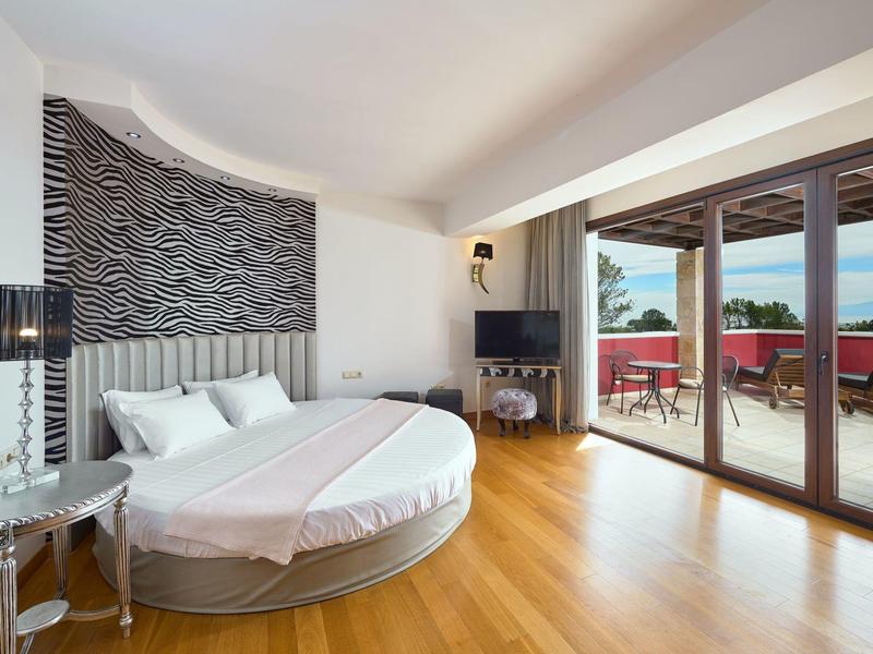 Modernes, rundes Bett in hellem Schlafzimmer mit Holzfußboden und Zugang zu roter Terrasse mit Meerblick.