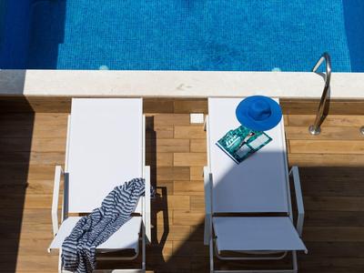 Zwei weiße Liegestühle neben blauem Pool, einer mit blauem Hut und Buch, Holzterrasse.