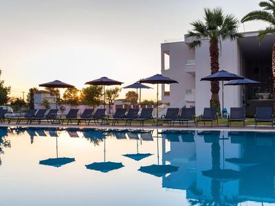 Pool mit blauer Wasserfläche, Liegestühlen, Sonnenschirmen und Palmen vor modernem Gebäude.