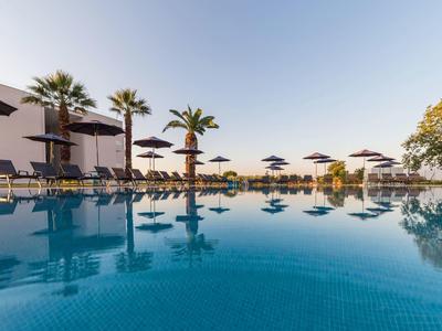 Blauer Pool mit Sonnenliegen, Schirmen, Palmen und klarstem Himmel im Hintergrund.