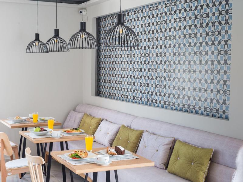 Modernes Café mit hellen Holztischen, grauen Stühlen, gepolsterter Bank und gemustertem Wandbild.