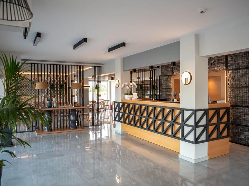 Heller Hotel-Empfang mit weißem Tresen, Holzverkleidung, Pflanzen und moderner Beleuchtung.