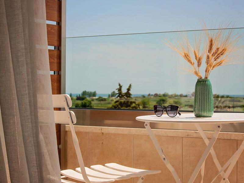 Balkon mit hellem Holzstuhl, weißem Tisch, grüner Vase mit Trockenblumen und Sonnenbrille vor Landschaft.