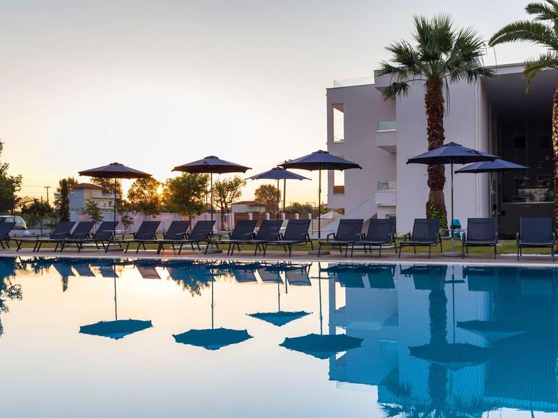 Pool mit blauer Wasserfläche, Liegestühlen, Sonnenschirmen und Palmen vor modernem Gebäude.