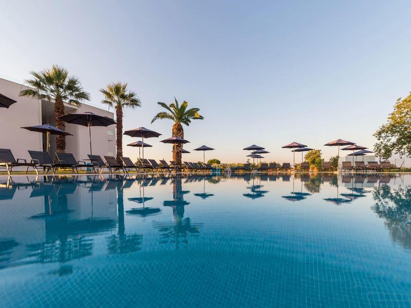 Blauer Pool mit Sonnenliegen, Schirmen, Palmen und klarstem Himmel im Hintergrund.