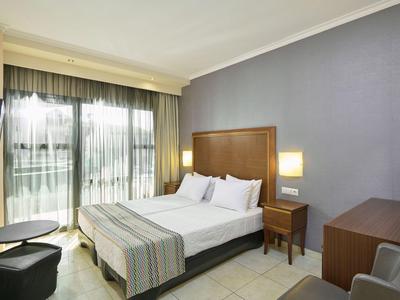 Camera d'hotel con letto matrimoniale, comodini, scrivania e balcone con tende trasparenti.