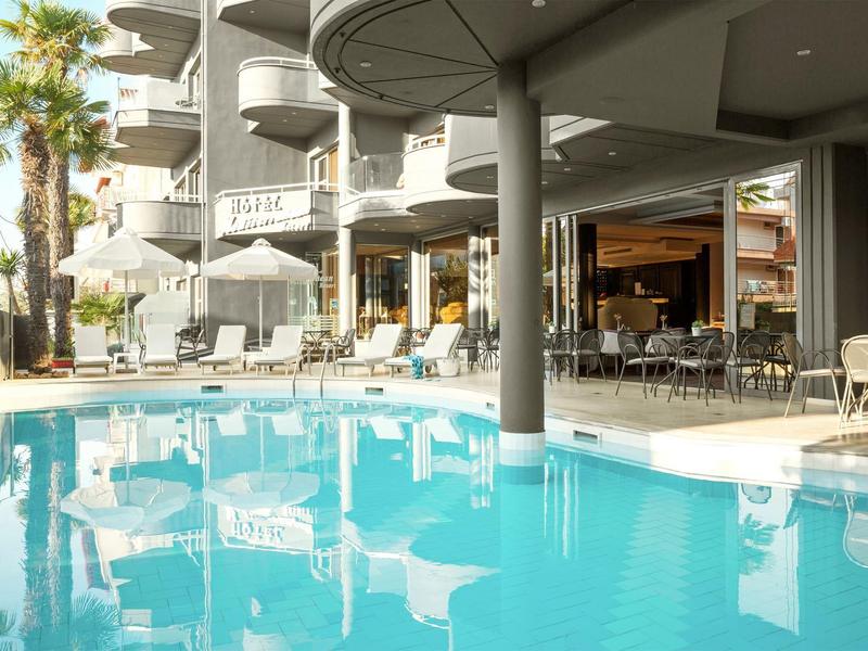 Moderní hotelový bazén s židlemi, stoly a zakřivenými balkony pod jemným světlem.