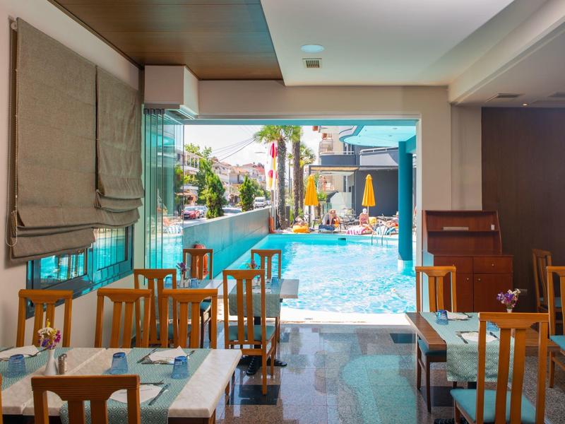 Vnitřní jídelní prostor s dřevěnými židlemi a stoly s výhledem na slunný bazén se slunečníky.