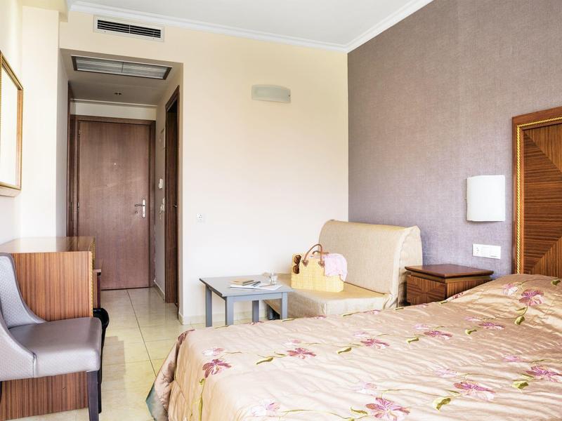 Hotelový pokoj s postelí, křeslem, malým stolem a dřevěnými dveřmi v pozadí.