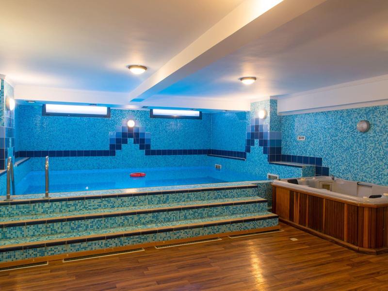 Vnitřní bazén se schody a vířivkou v místnosti s dřevěnou podlahou a modrými dlaždicemi.