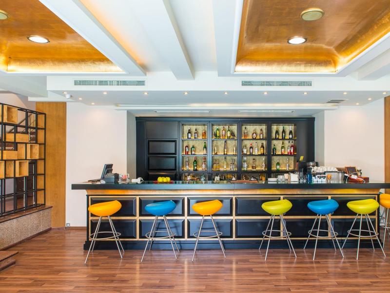 Moderní hotelový bar s barevnými stoličkami, dřevěnou podlahou a teplým osvětlením stropu.