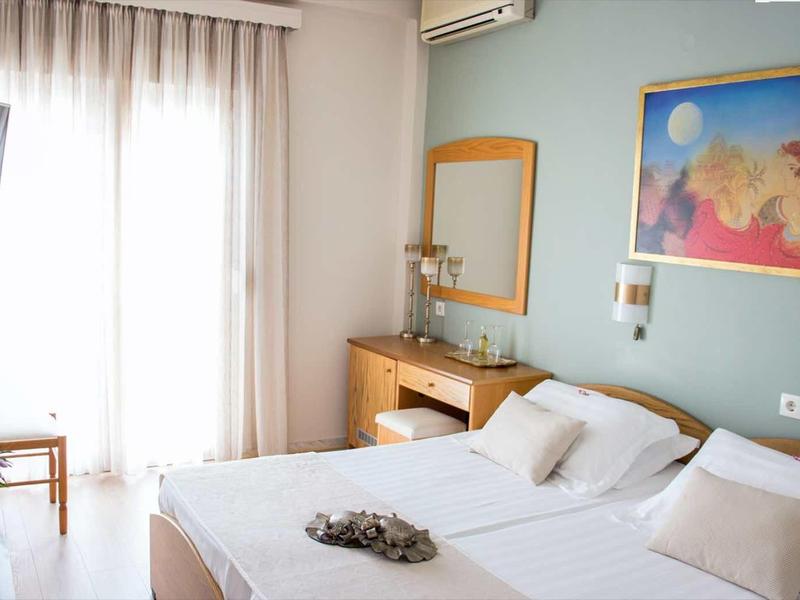 Světý hotelový pokoj s manželskou postelí, psacím stolem, židlí, televizí a balkonem se závěsy.