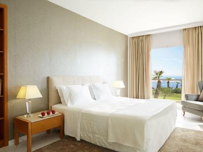 Jasny pokój hotelowy z białym łóżkiem podwójnym, lampkami nocnymi i widokiem na morze przez duże okno.