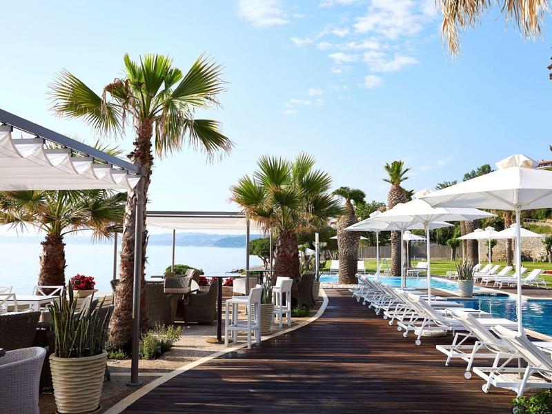 Strefa basenowa z leżakami, parasolami i palmami obok hotelu z widokiem na morze.
