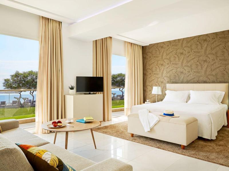 Nowoczesny pokój hotelowy z łóżkiem podwójnym, sofą, stołem i widokiem na ogród i morze.