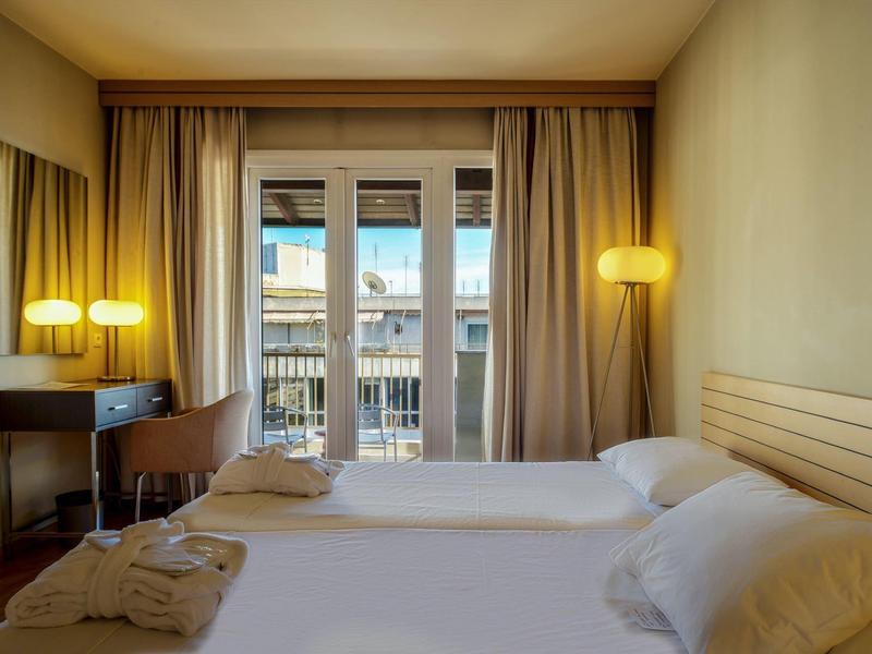 Camera d'albergo luminosa con due letti singoli, scrivania e vista su balcone e edifici.