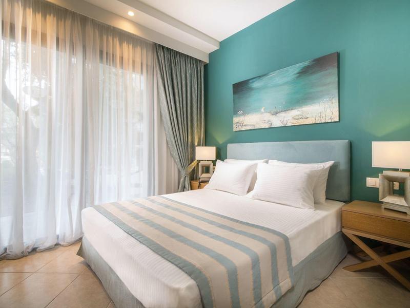 Camera d'hotel luminosa con letto matrimoniale, parete turchese e grandi finestre con tende.