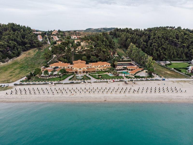 Ośrodek wypoczynkowy przy plaży z leżakami, zielonymi ogrodami i zalesionymi wzgórzami w tle.